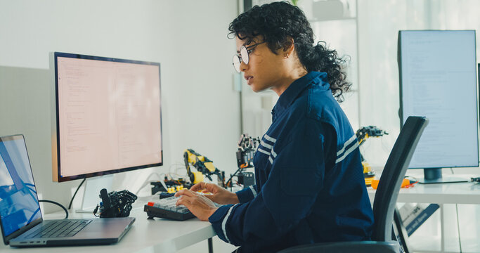 Young African American female robotics engineer coding AI software for robotic arm in laboratory. Embedded system, firmware testing, machine learning, industrial automation, future technology concept.