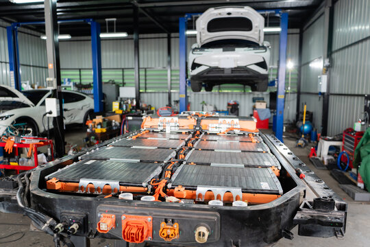 Inside an electric vehicle battery repair garage, an EV battery pack is checked beside cables, connectors, and a tool cart.