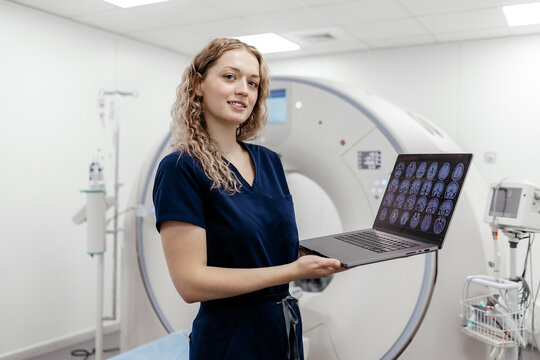 Smiling female medical professional presenting brain scans on a laptop beside a CT scanner, concept of brain MRI, CT imaging, radiology and modern healthcare.