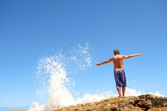 Barefoot fisherman bracing against crashing wave on rocks