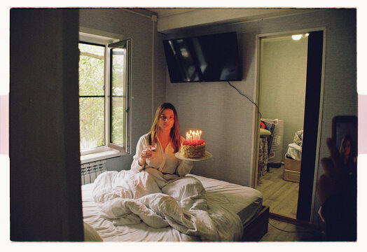 A woman holds a birthday cake with lit candles alone in bed