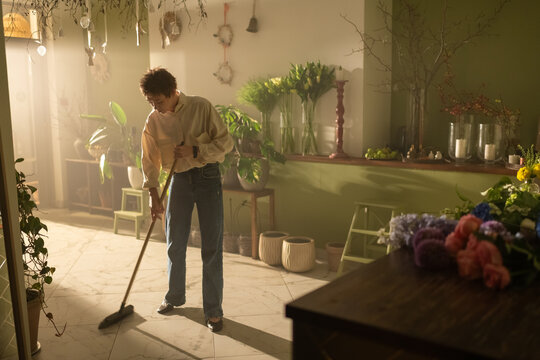 Young female florist sweeps floor with broom in closed flower shop