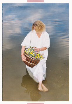 A woman wades barefoot into still water carrying wildflowers