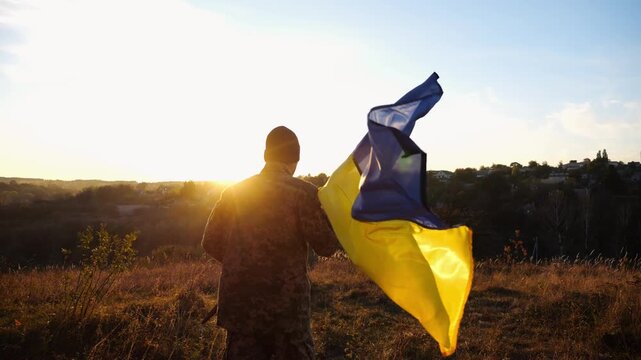 Young man in military uniform jogs with waving flag of Ukraine at sunset. Male soldier of ukrainian army running with lifted blue-yellow banner at countryside. Concept of invasion resistance