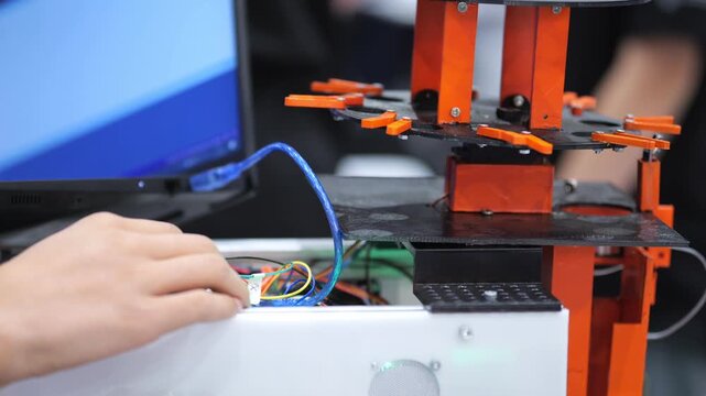 Hand adjusts wires near 3D printer. Orange print head moves above white build plate. Blue cable connects printer to computer. Background shows blurred monitor and workspace. Focus on mechanical action