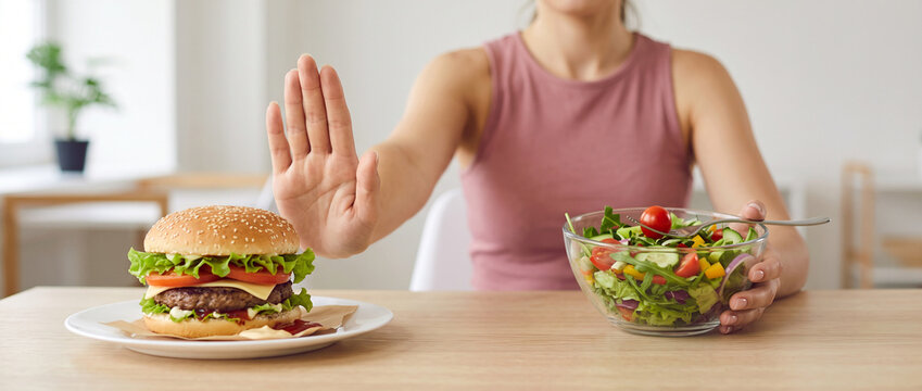 Person choosing a fresh vegetable salad instead of a high-calorie burger, showing a stop gesture toward junk food while following a healthier diet for weight control, healthy diet, fast food refusal, 