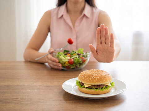 Woman refusing a hamburger while holding a bowl of fresh salad, raising her open palm to reject high-calorie fast food during a weight loss diet, healthy eating, junk food avoidance, calorie control, 