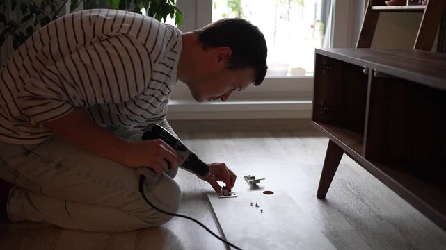 Concentrated man on his knees assembling flat pack furniture, using an electric drill to carefully screw a metal hinge onto a wooden cabinet door, illustrating a diy home improvement project