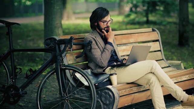 Businessman Working On Laptop Sitting On Wooden Bench Next To Bicycle In Park. Remote Work 