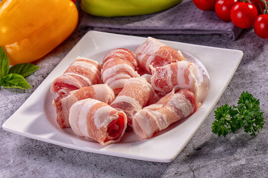 Raw bacon wrapped meat rolls on white square plate with yellow and green bell peppers, cherry tomatoes, and fresh herbs on gray stone surface