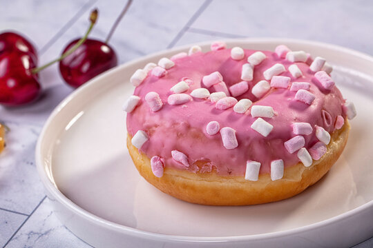 Pink glazed donut topped with mini marshmallows on white plate, fresh cherries in background, soft lighting and shallow depth of field
