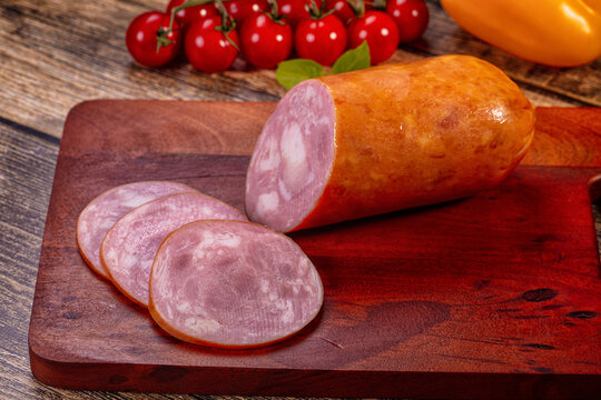 Sliced ham sausage on dark wooden cutting board with cherry tomatoes and yellow pepper in background, rustic food presentation