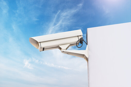 White outdoor CCTV camera monitors territory from building wall corner under blue cloudy sky. Modern electronic surveillance