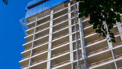Unfinished concrete high-rise building under construction with safety netting and construction elevator against clear blue sky. © sommersby