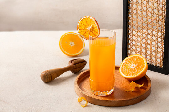 Fresh orange juice in a tall glass with dried orange garnish on a wooden tray, with halved oranges, wooden citrus juicer, orange peel, and rattan frame
