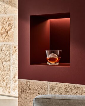 Whiskey glass with ice sphere in recessed maroon niche interior still life with stone wall for design card poster background