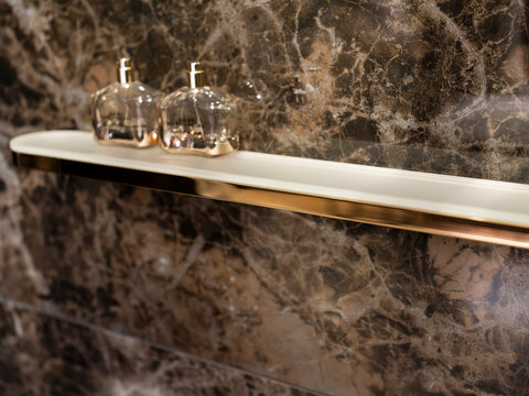 Two glass flacons with gold pumps on a small cream shelf with copper edge, set against veined dark brown marble.