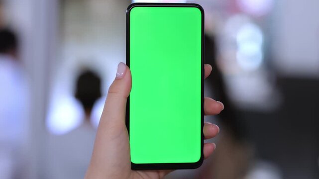 Hand holds smartphone with green screen. Background blurred, people visible. Phone held steady in three keyframes