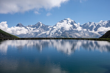 Fototapeta premium Dramatic snow-covered mountain range mirrored in calm blue alpine water beneath a clear summer sky.