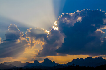 Dramatic tropical sunset over the seaside, with golden sun rays breaking through layers of clouds and illuminating the sky above the calm ocean. The warm evening light creates a striking atmosphere, w © Yuliia