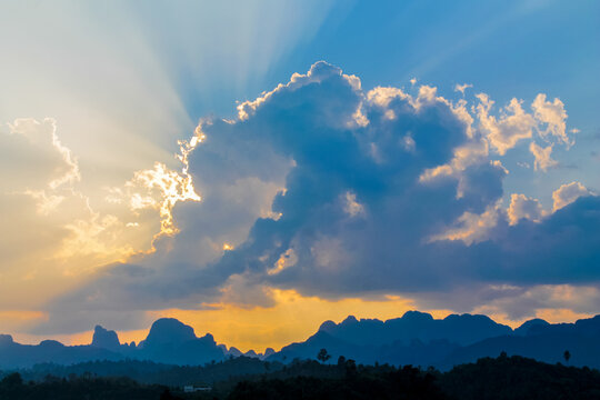 Dramatic tropical sunset over the seaside, with golden sun rays breaking through layers of clouds and illuminating the sky above the calm ocean. The warm evening light creates a striking atmosphere, w