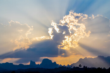 Dramatic tropical sunset over the seaside, with golden sun rays breaking through layers of clouds and illuminating the sky above the calm ocean. The warm evening light creates a striking atmosphere, w © Yuliia