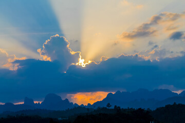 Dramatic tropical sunset over the seaside, with golden sun rays breaking through layers of clouds and illuminating the sky above the calm ocean. The warm evening light creates a striking atmosphere, w © Yuliia