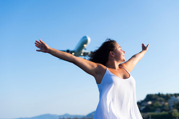 A woman in a white dress stands outdoors with her arms outstretched as an airplane flies overhead in Kerkira, Greece. The clear blue sky and coastal landscape frame the background. © dsbedarieva
