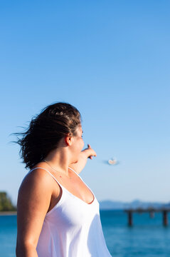 A woman in a white dress points toward an approaching airplane over the sea in Kerkira, Greece. The coastal setting features a clear blue sky and a pier in the background.