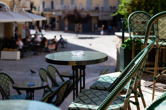 An outdoor cafe terrace features bistro tables and woven green chairs in Kerkira, Greece. A clear glass ashtray rests on the small round table overlooking an open city square.