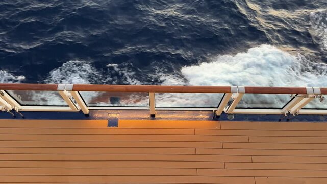 Top view of a wooden cruise ship deck and glass railing with white ocean waves churning below. Luxury travel concept, peaceful summer vacation, tropical marine voyage, ocean cruise