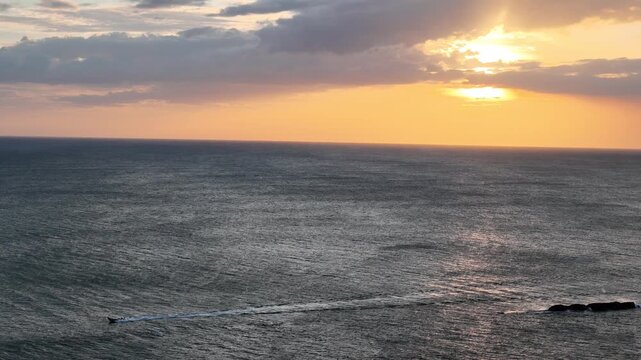 A boat moves through the ocean as the sun sets on the horizon. The sky shows warm colors while waves roll in.