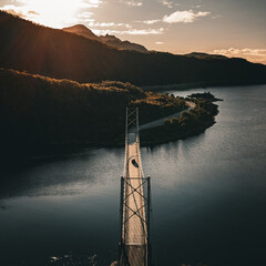 Aerial vertical view of a suspension bridge over a fjord with mountains and road during a bright sunset in Norway © Quality by Simon