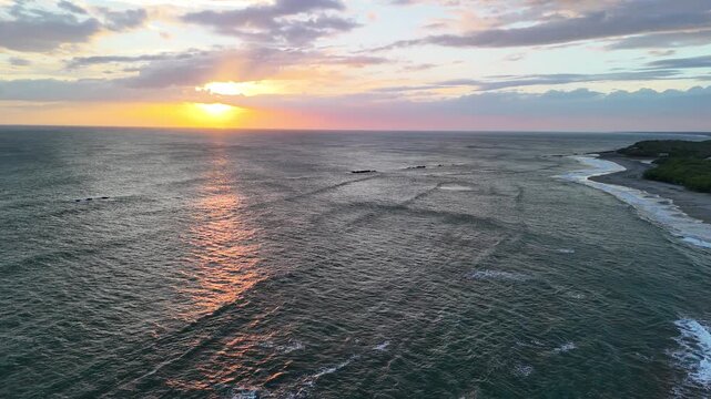 Waves roll onto the shore as the sun sets over the ocean near a beach. Clouds scatter the sky with colors from the fading light.