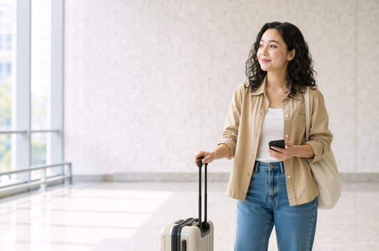 Young woman with suitcase and smartphone waiting in airport terminal. Vertical portrait of female traveler holding luggage in modern transit station. Vacation and journey concept