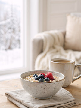 Bowl of oatmeal with fresh berries and mug of coffee on wooden table. Vertical photo of healthy winter breakfast by window with snowy background. Cozy morning concept