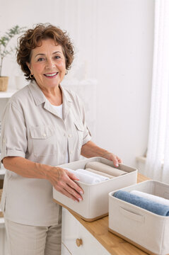 Smiling senior woman organizing folded towels in storage bins at home. Vertical portrait of happy mature female doing domestic chores and tidying up. Copy space for text