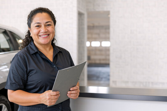 Smiling female auto service advisor holding clipboard. Middle aged hispanic woman working at car dealership counter. Customer service concept. Copy space for text
