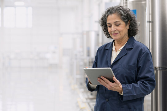 Mature female engineer using tablet in manufacturing plant. Vertical portrait of smiling woman in blue uniform inspecting industrial production. Copy space for text