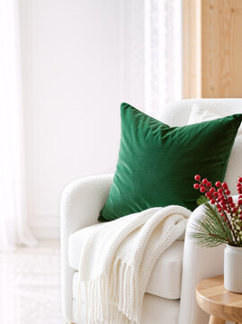 White armchair with green velvet pillow and knit blanket in bright living room. Cozy winter home interior with holiday decor and pine branches. Copy space for text