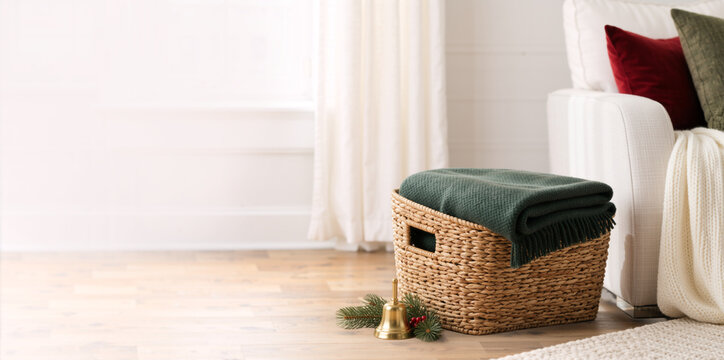 Woven basket with green blanket and brass bell on wooden floor. Cozy winter living room interior with decorated white sofa. Copy space for text