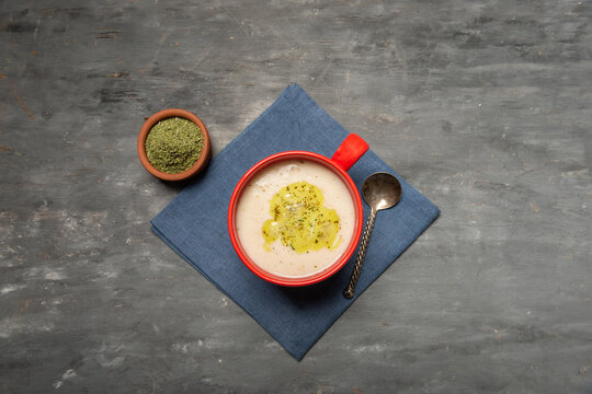 Traditional Turkish yogurt soup served in a red ceramic cup, garnished with melted butter and dried mint. Overhead food composition with a blue napkin and generous copy space on a rustic dark table.