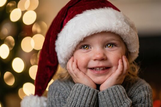 Cute little girl wearing Santa hat smiling with head in hands near Christmas tree lights