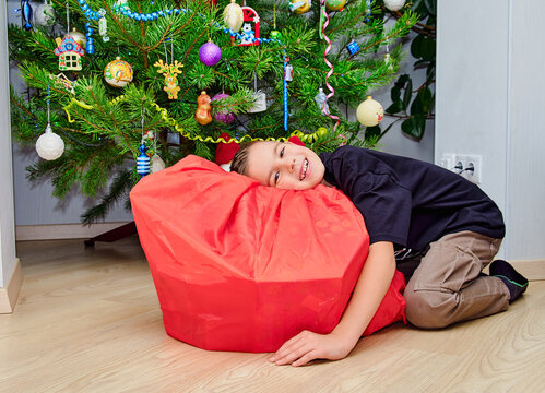 Happy child lying beside a large Christmas gift in front of a decorated Christmas tree, celebrating the holiday season at home with a joyful festive atmosphere
