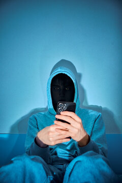 Hooded person using a smartphone in dramatic blue lighting, creating a mysterious cybercrime and online privacy concept with strong shadows and a dark, technological atmosphere