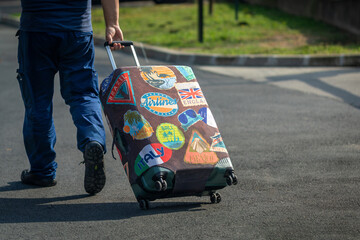 Rear view of an unrecognizable traveler walking with a wheeled suitcase covered in colorful travel themed stickers on an asphalt road, symbolizing vacation, tourism, travel and journey. © Studio Aurelia