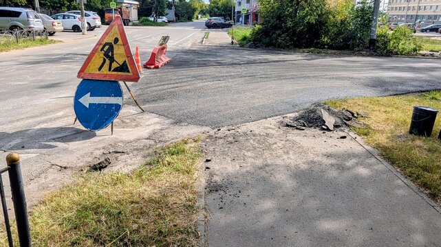 city street construction site with temporary road signs indicating road works and detour direction, fresh black asphalt patch, and safety cones under bright 
