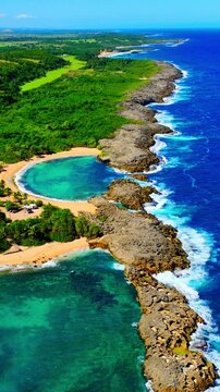 Aerial view of Mar Chiquita tidal pool and rocky coast, Puerto Rico. Circular turquoise lagoon beside golden sand, dense green forest and deep blue ocean with white surf. Vertical video.