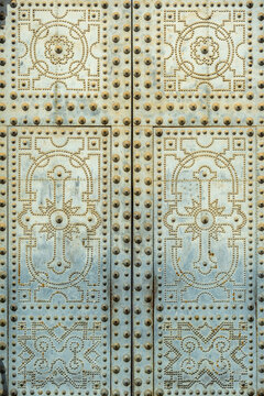 Antique metal door with intricate repeating geometric and floral patterns studded with round rivets, weathered textured surface evoking vintage, rustic architectural detail