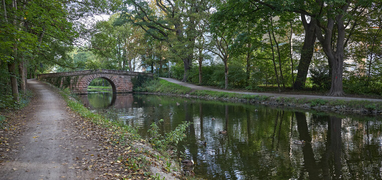 Stone arch bridge over a calm canal surrounded by lush green trees in a peaceful park setting